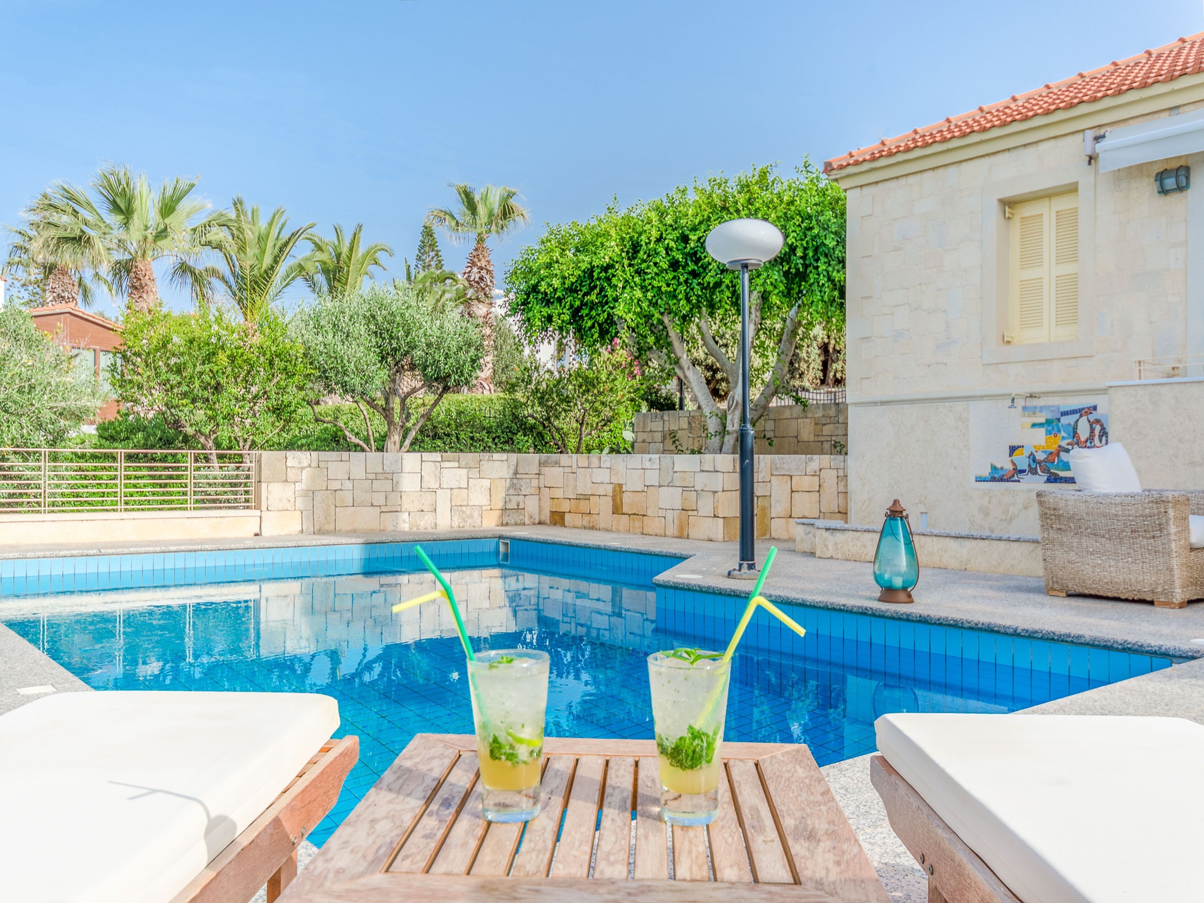 A pool area with two lounge chairs, a table with drinks, and a building with a tiled roof.