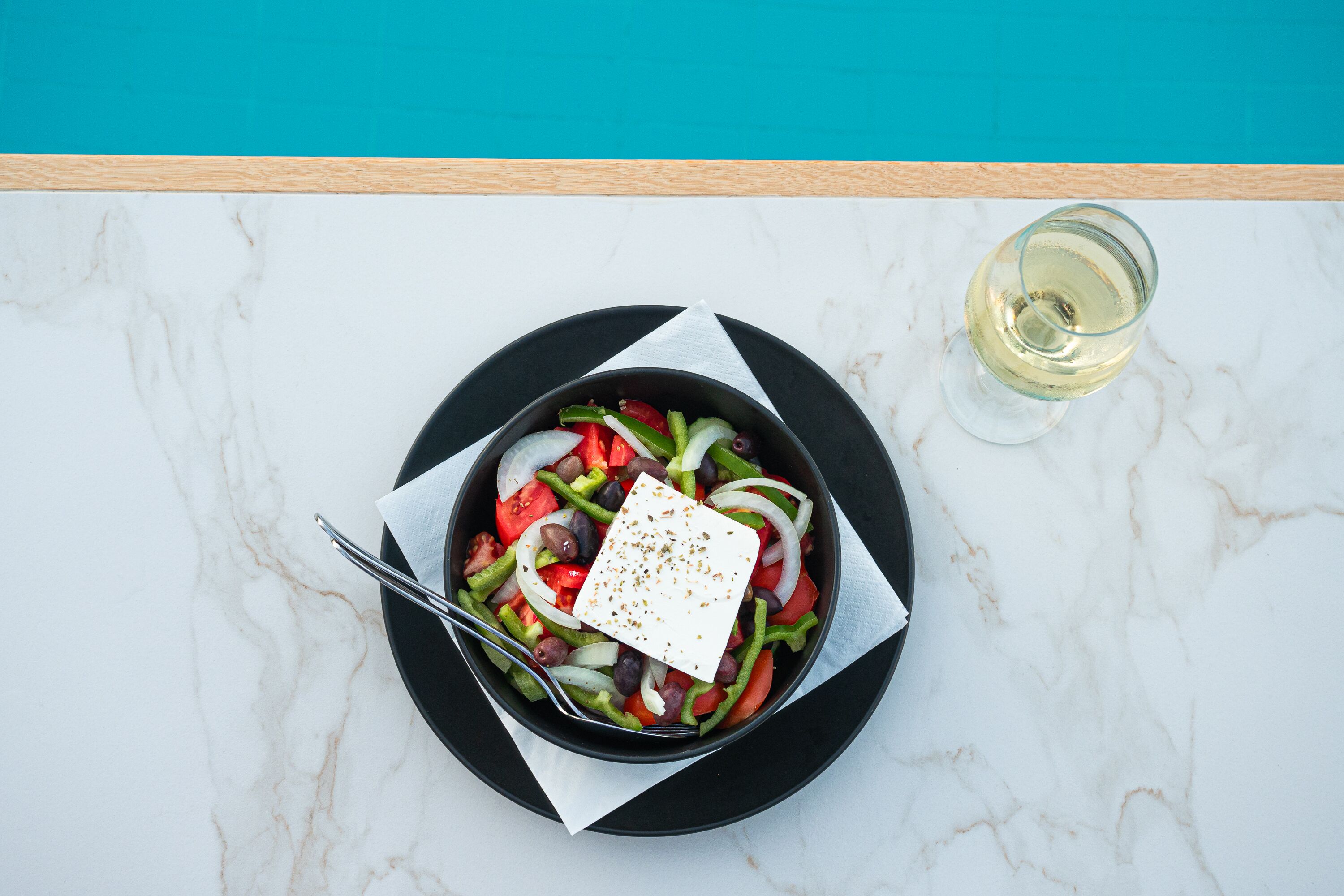 A salad on a black plate with a white napkin, a fork, and a glass of white wine on a marble surface.
