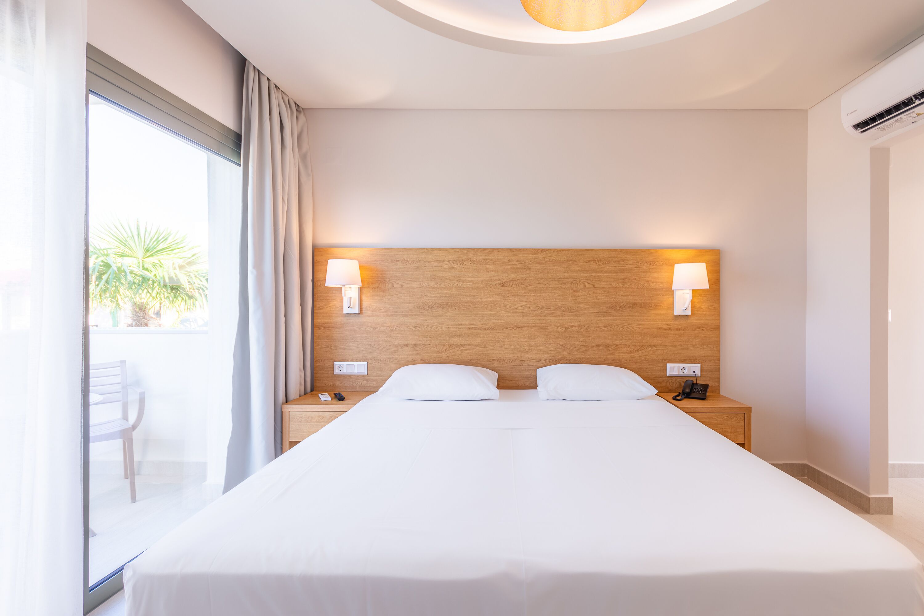 Habitación de hotel con una cama grande, dos mesitas de noche, un cabecero de madera y vistas al exterior.