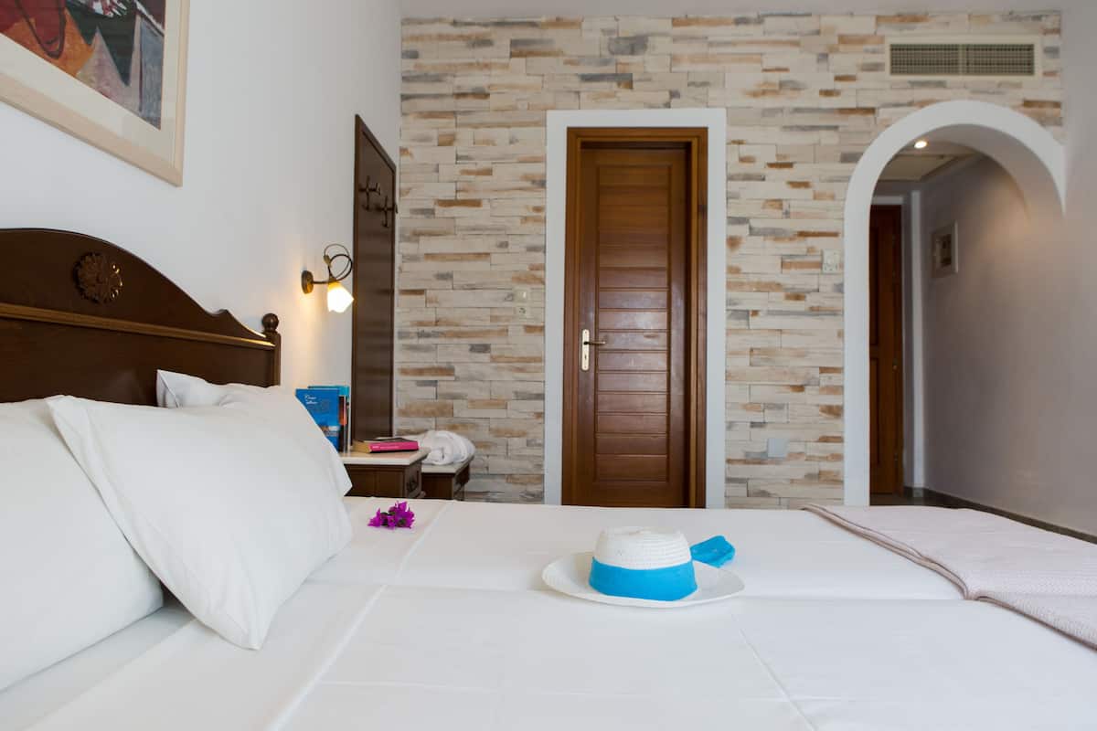 A hotel room with a stone wall, a wooden door, a bed with white bedding, and a small table with a book and a pink object.