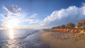 Una playa con hileras de tumbonas naranjas y sombrillas de paja, un cielo despejado y el sol poniéndose sobre el océano.