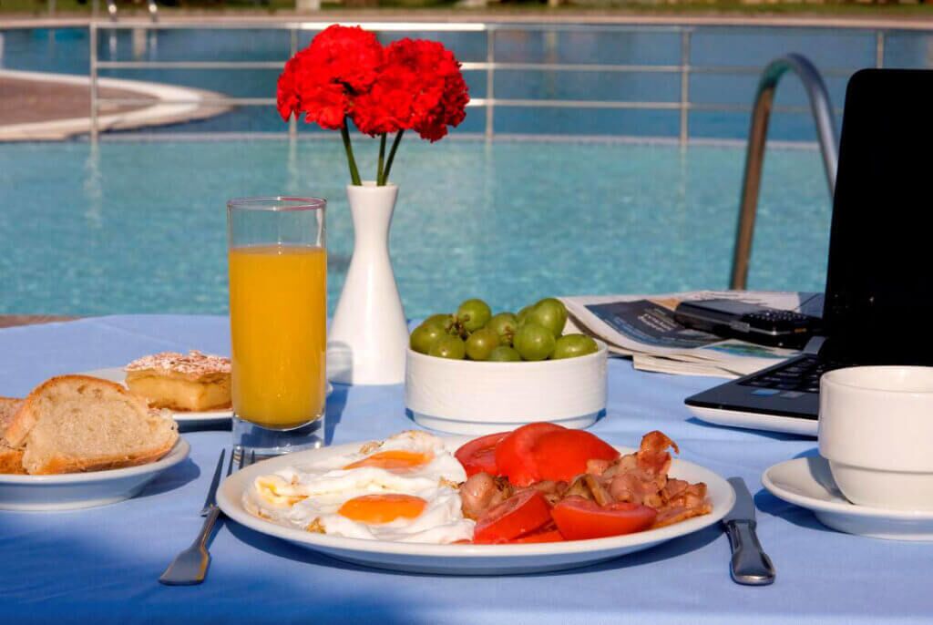 A breakfast setup by a pool with eggs, bacon, tomatoes, toast, and a glass of orange juice.