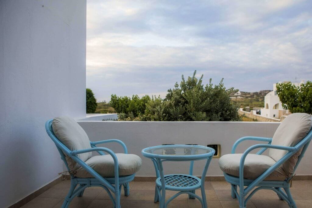Ein Balkon mit zwei Korbstühlen und einem kleinen Tisch, mit Blick auf eine Landschaft mit Grünflächen und weißen Gebäuden.