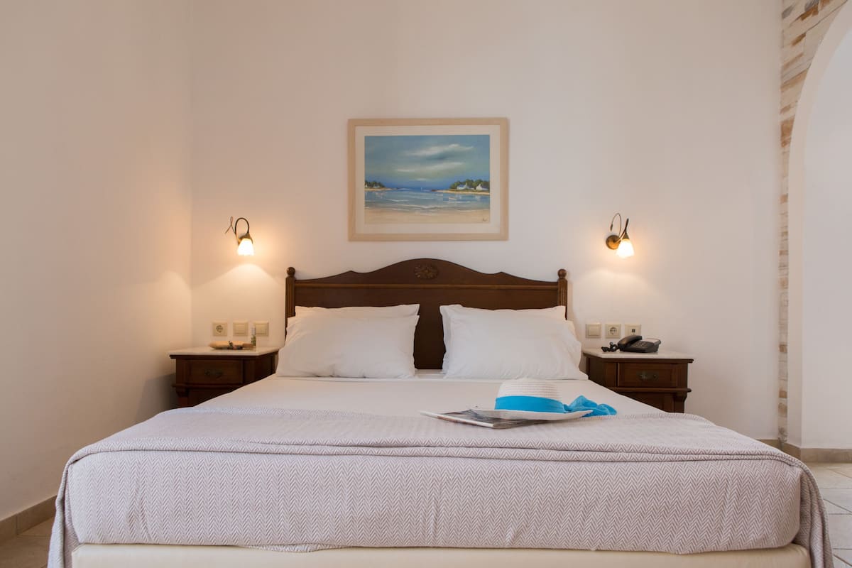A neatly made bed with a white and beige patterned bedspread, two bedside tables with lamps, a framed painting of a beach scene, and a wooden headboard.