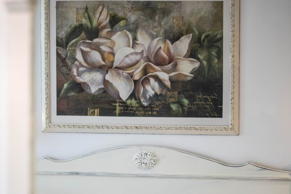 A bedroom with a bed, white pillows, and a framed painting of flowers on the wall.