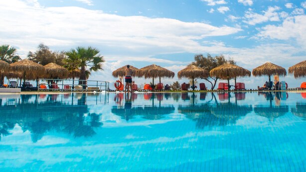 Una piscina con agua cristalina azul, rodeada de sillas de descanso rojas y sombrillas de paja.