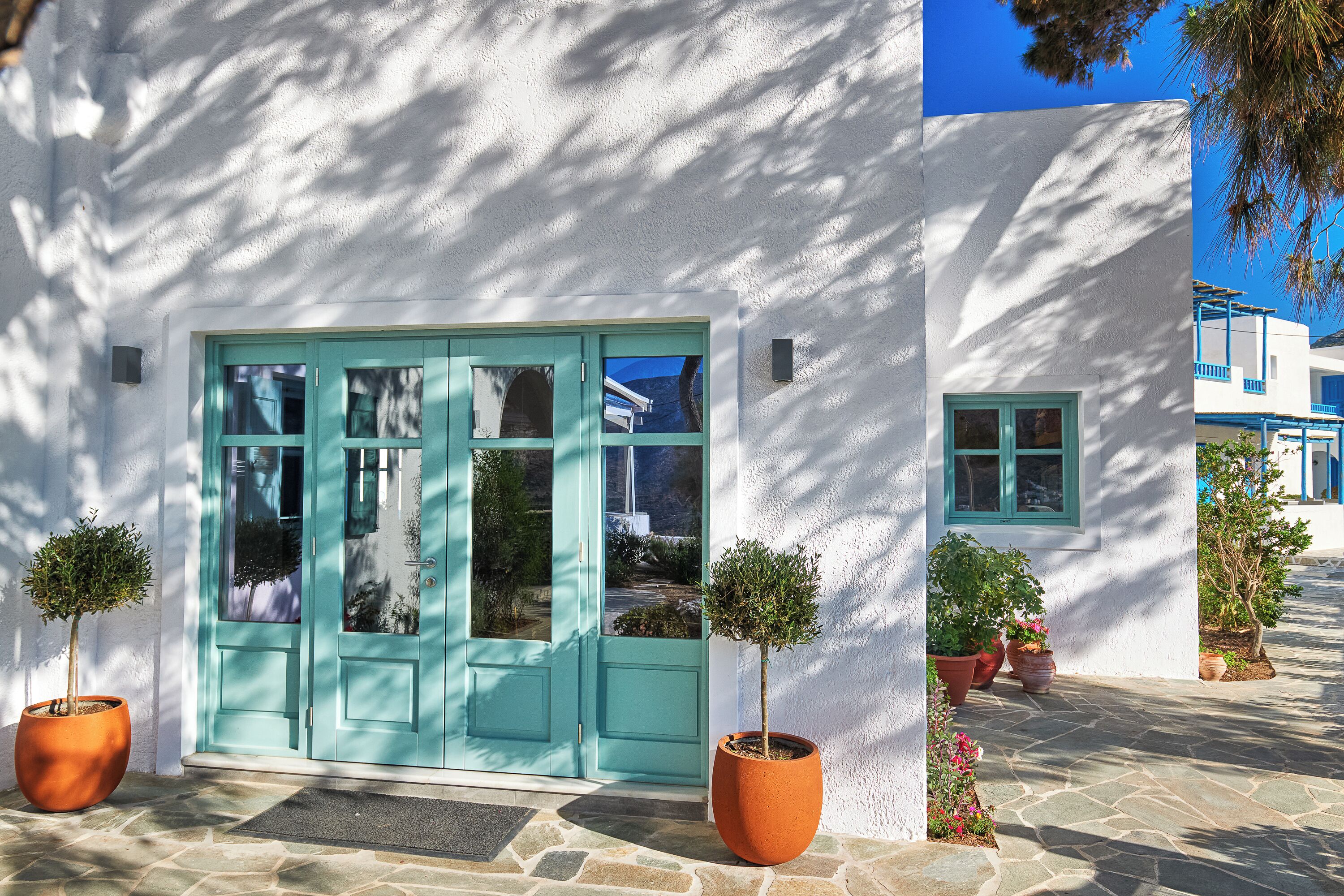 A white building with a turquoise door and windows, potted plants, and a stone-paved entrance.
