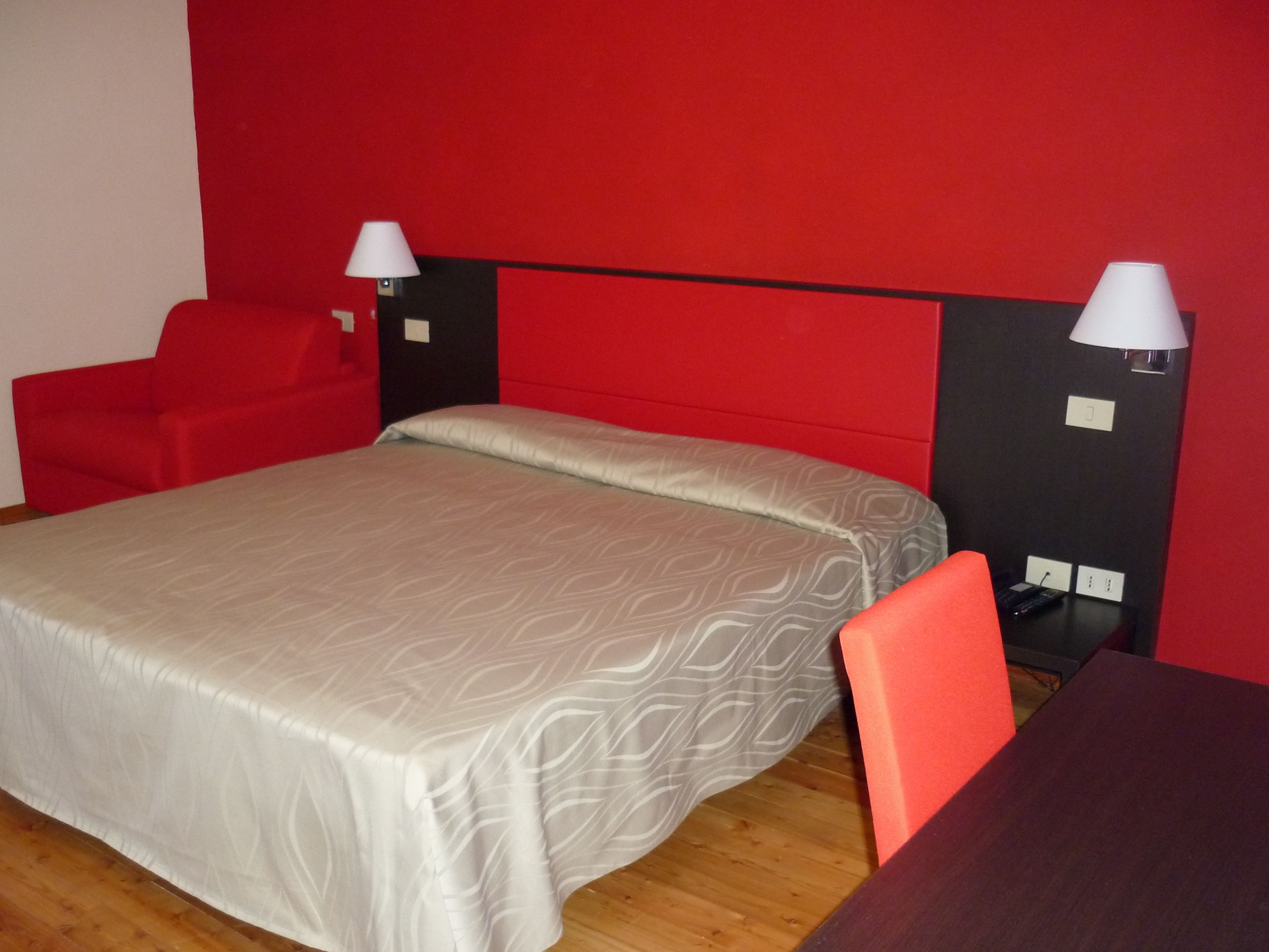 Una habitación de hotel con cama, una silla roja, una mesa de madera y dos lámparas de noche.