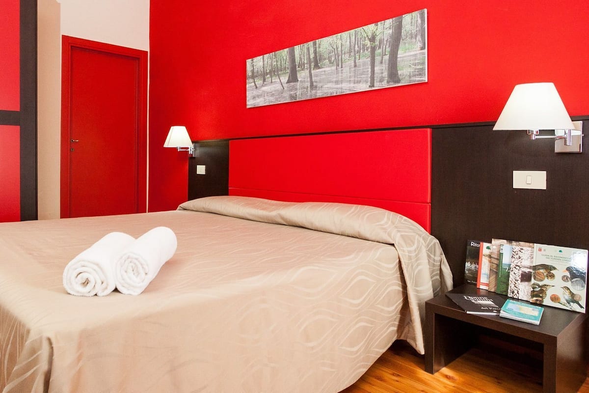 A hotel room with a red wall, a bed with beige bedding, two bedside lamps, a nightstand with books, and a door with a red frame.