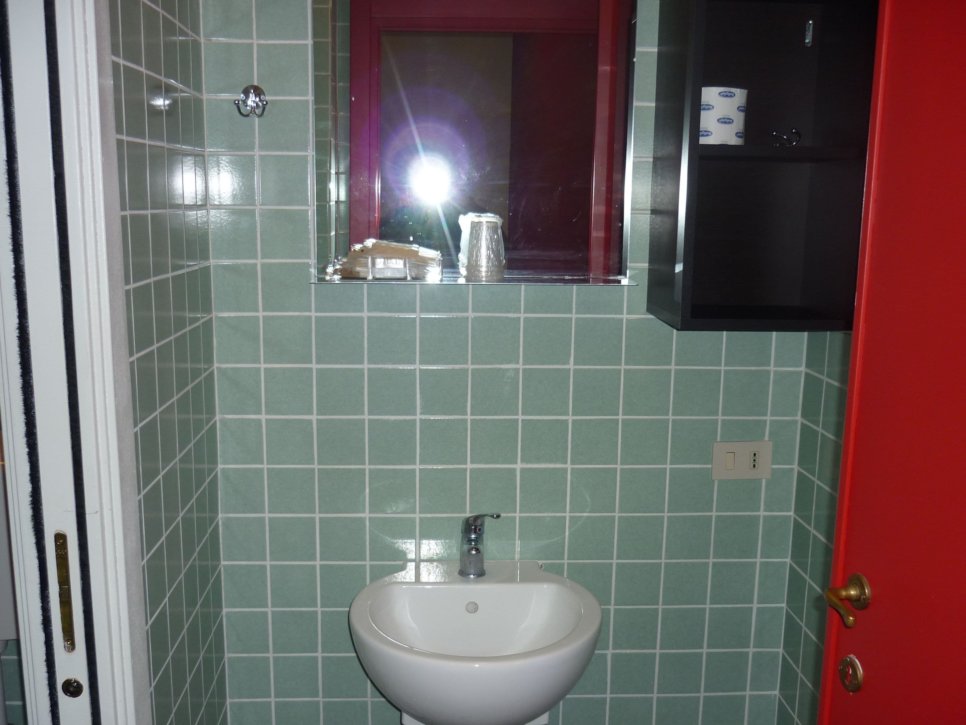 Een badkamer met een witte wastafel, groene betegelde wanden, een spiegel en een rode deur.