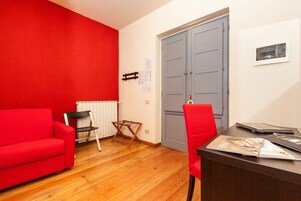 Una habitación con un sofá rojo, una silla de madera, un escritorio con papeles, un radiador y una puerta gris.