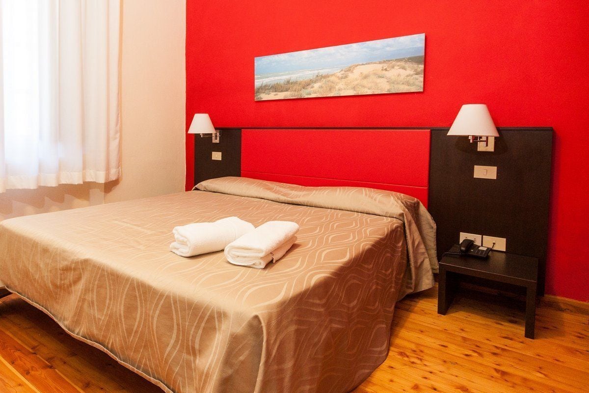 Une chambre d’hôtel avec un mur rouge, un lit recouvert d’une couvre-lit à motifs beige, deux tables de chevet avec des lampes et un tableau représentant une scène de plage.