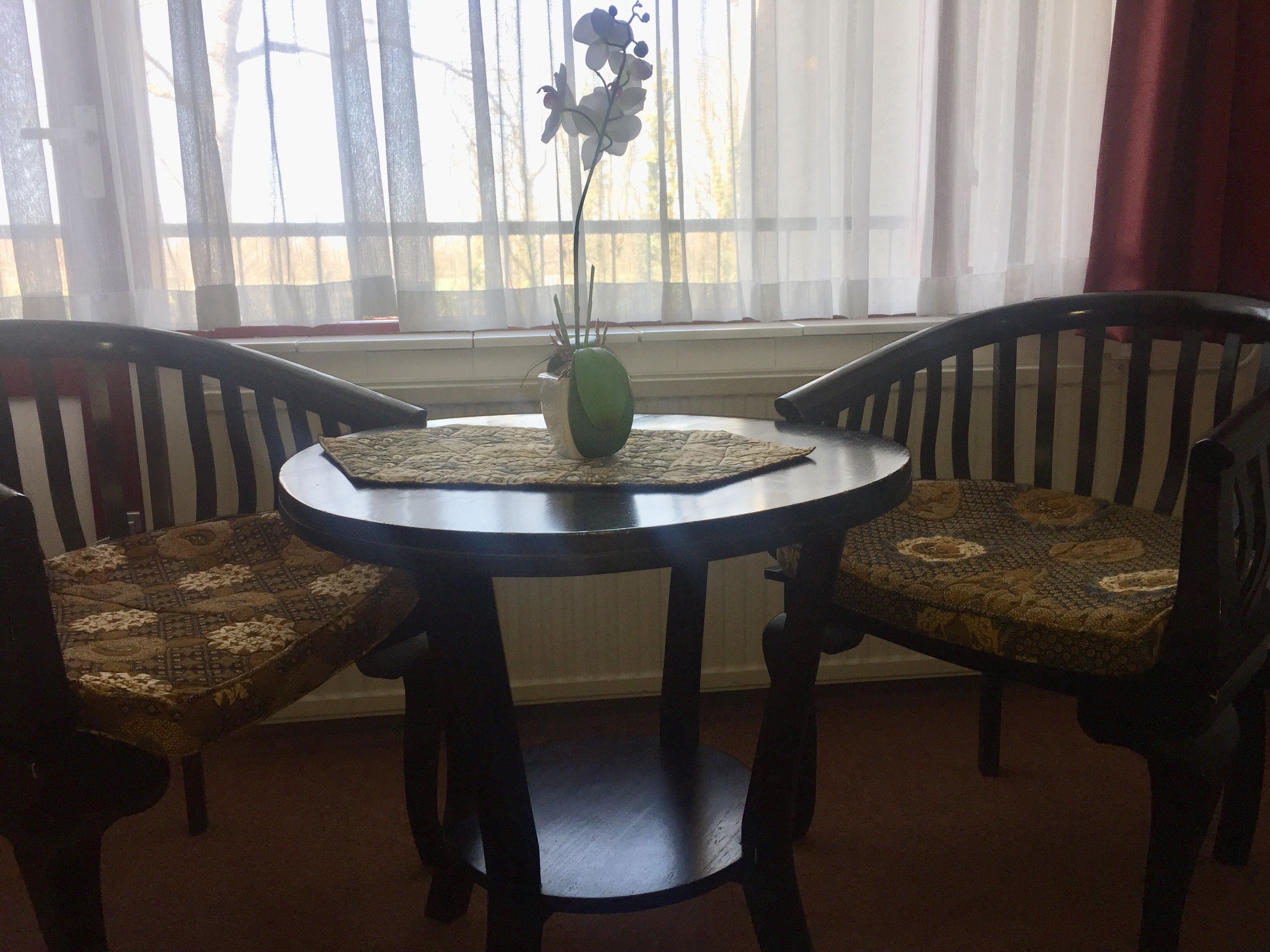 Une table ronde surmontée d’un set de table en fibres tressées, deux chaises avec des coussins à motifs et un vase vert contenant une plante.