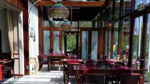 A restaurant with wooden tables and chairs, red tablecloths, and large windows offering views of the outdoors. - Hotel Den Helder (Den Helder)