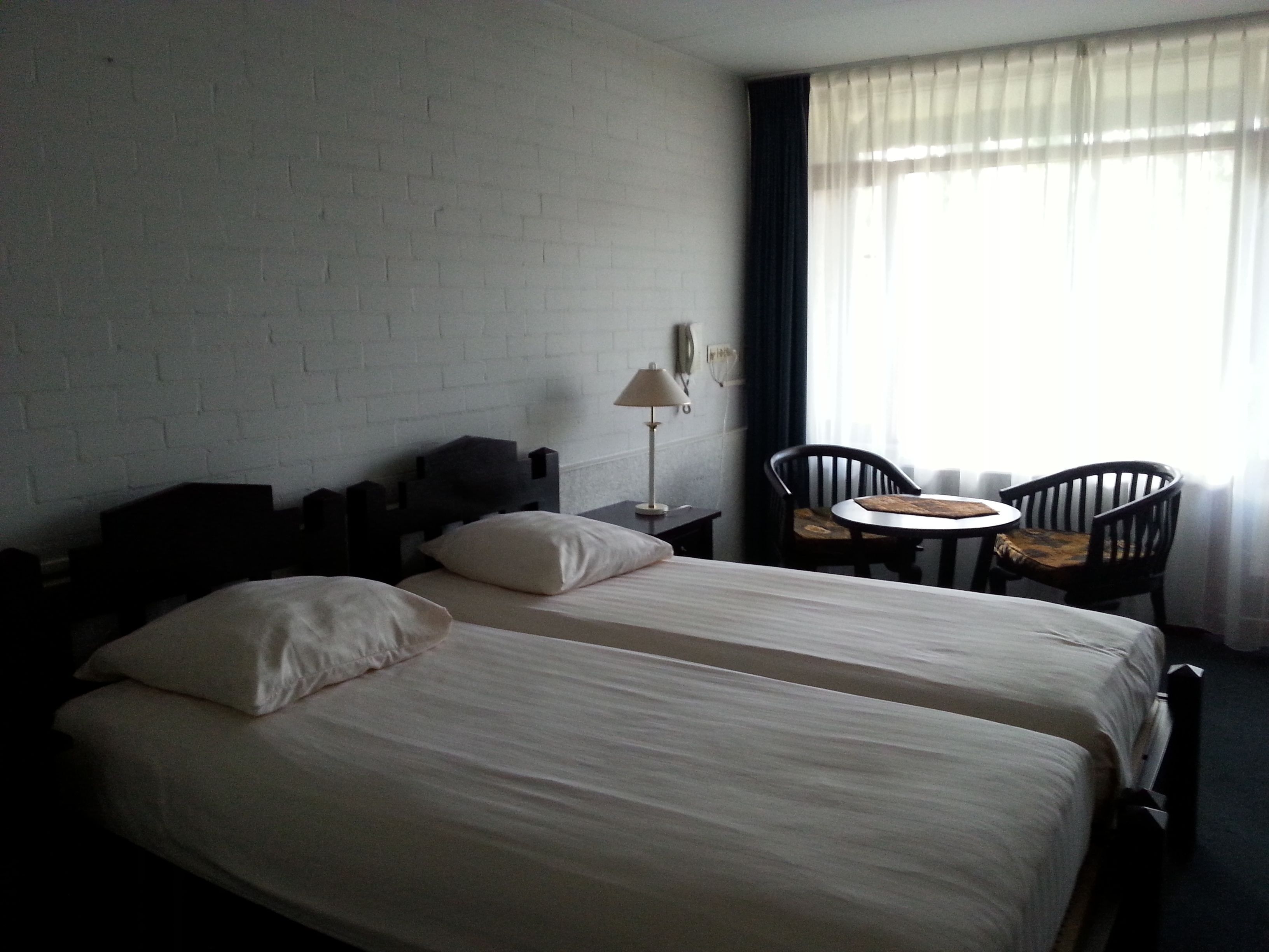 Une chambre d’hôtel avec deux lits, une petite table, deux chaises, une lampe et une fenêtre avec des rideaux.