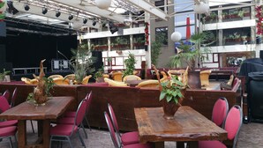An outdoor seating area with wooden tables and red cushioned chairs, surrounded by potted plants and a stage with lighting equipment. - Hotel Den Helder (Den Helder)
