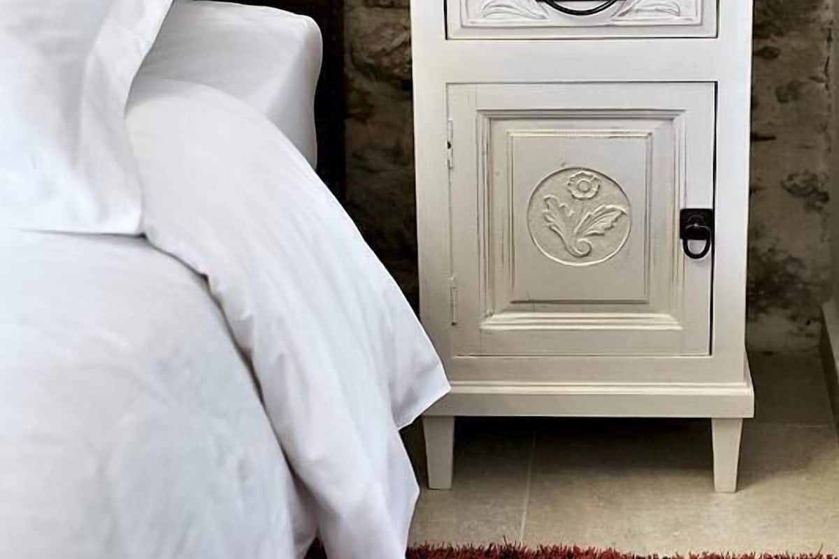 A bed with white linens, a brown headboard, and a white nightstand with a vase and a small lamp.
