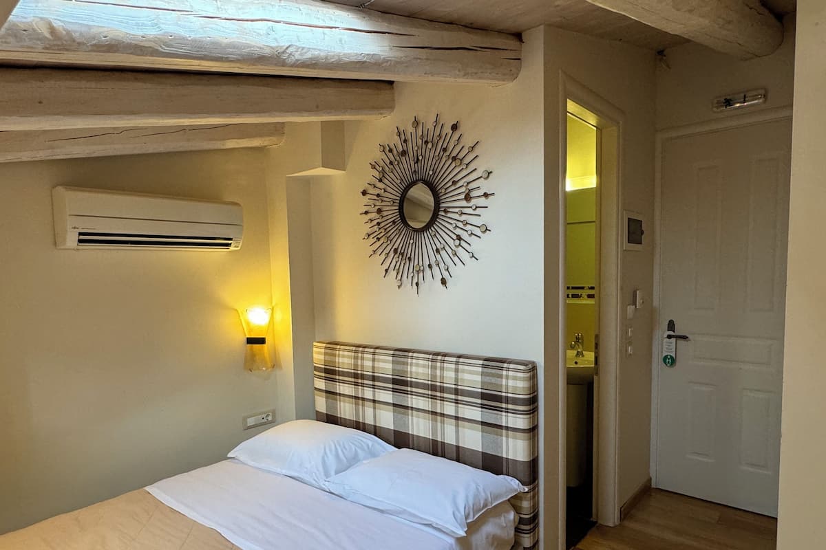 A bedroom with a bed, a wall-mounted mirror, a wall light, and a door leading to a bathroom.