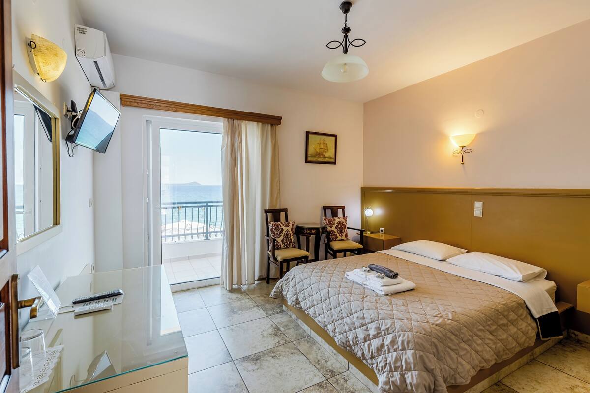 A hotel room with a bed, a desk with a computer, a chair, a table, a TV, and a view of the sea.
