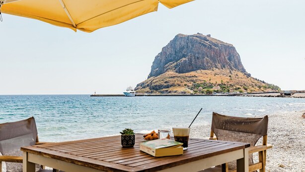 Una mesa de madera con un libro, un vaso de bebida y una planta pequeña bajo un paraguas amarillo en una playa con una colina rocosa al fondo.