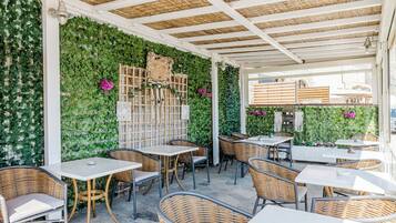 Café al aire libre con mobiliario de mimbre, mesas blancas y paredes cubiertas de follaje verde.