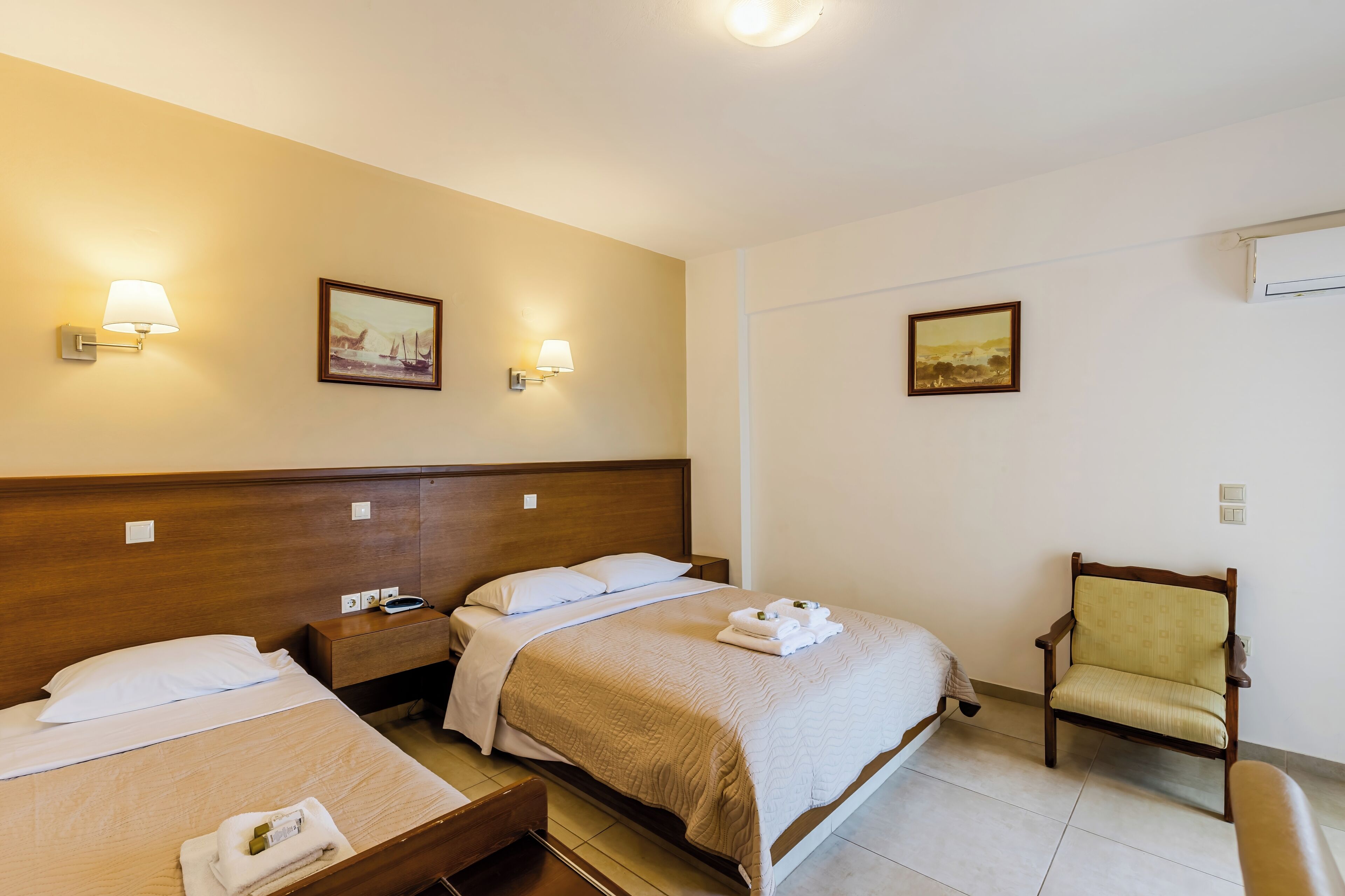 A hotel room with two beds, a chair, and wall-mounted lamps.