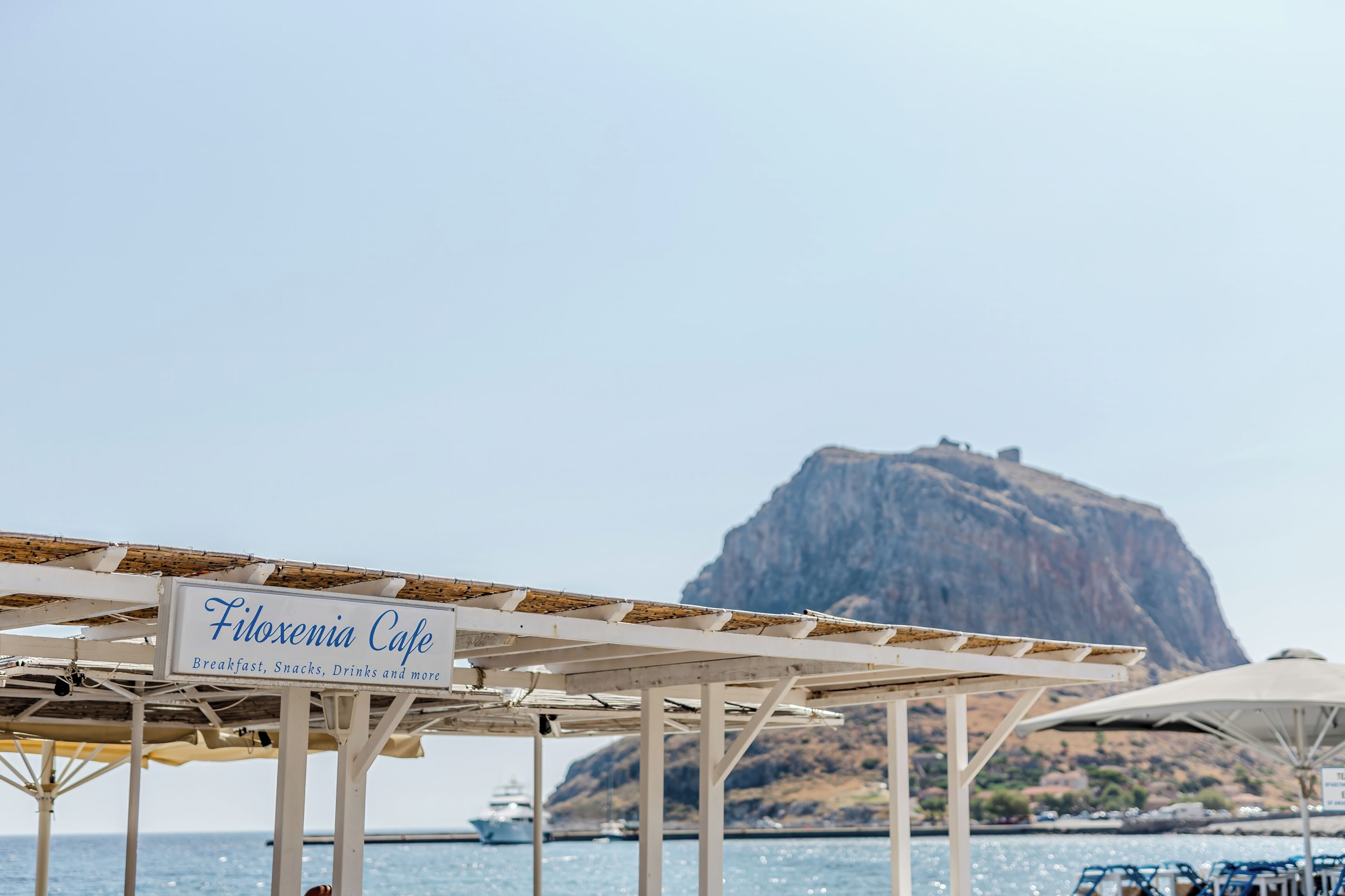 A beachside cafe named Filoxenia Cafe with a signboard, umbrellas, and a view of a large rock formation in the background.