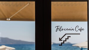 Una ventana con vistas al mar y a las montañas, y el texto 'Filoxenia Cafe' con una flecha señalando las escaleras.