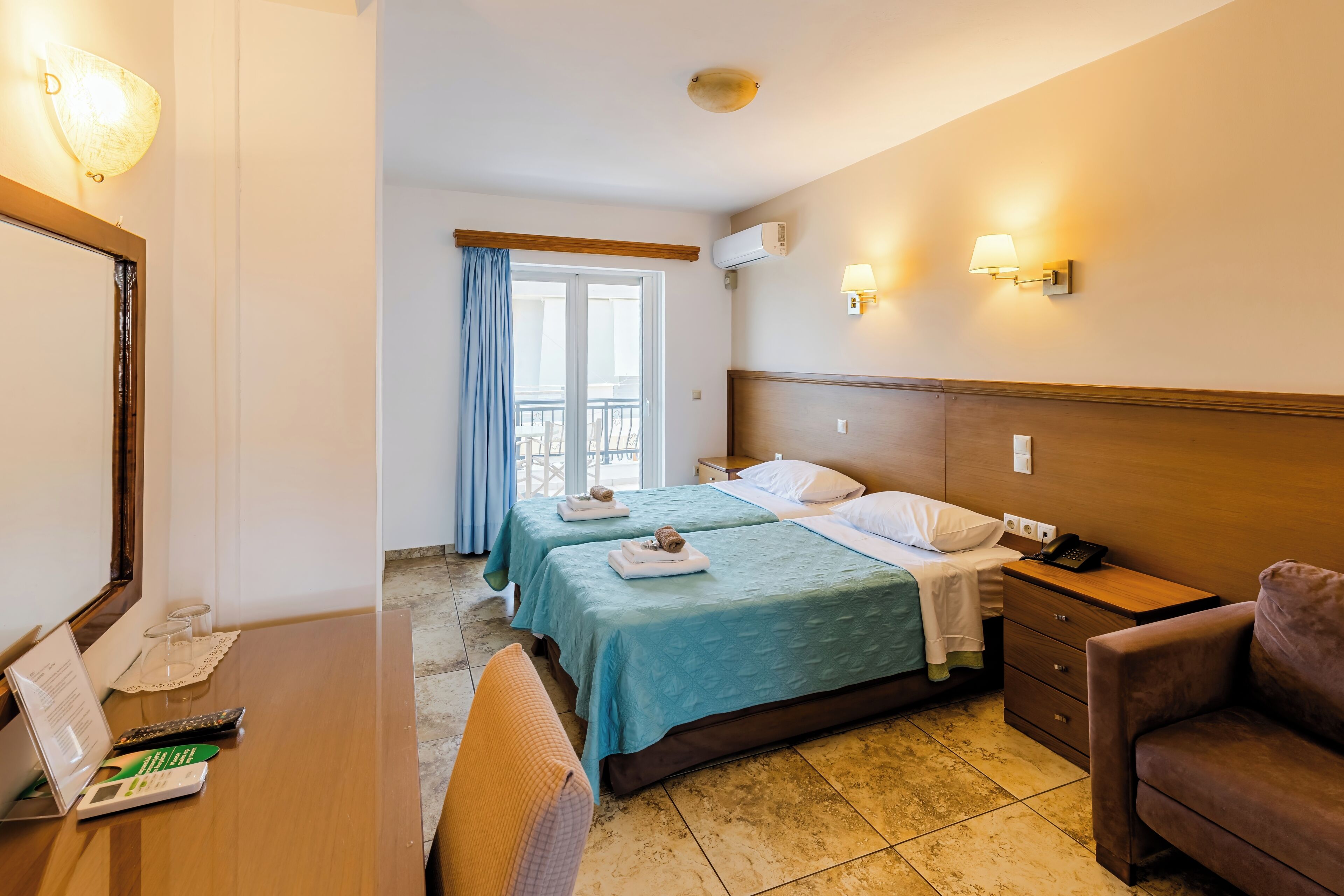 A hotel room with two beds, a desk with a chair, a small sofa, a balcony with a view, and a bathroom with a mirror and towel rack.