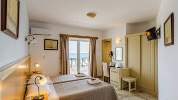 A hotel room with two beds, a desk, a chair, a TV, and a balcony with a view of the sea.