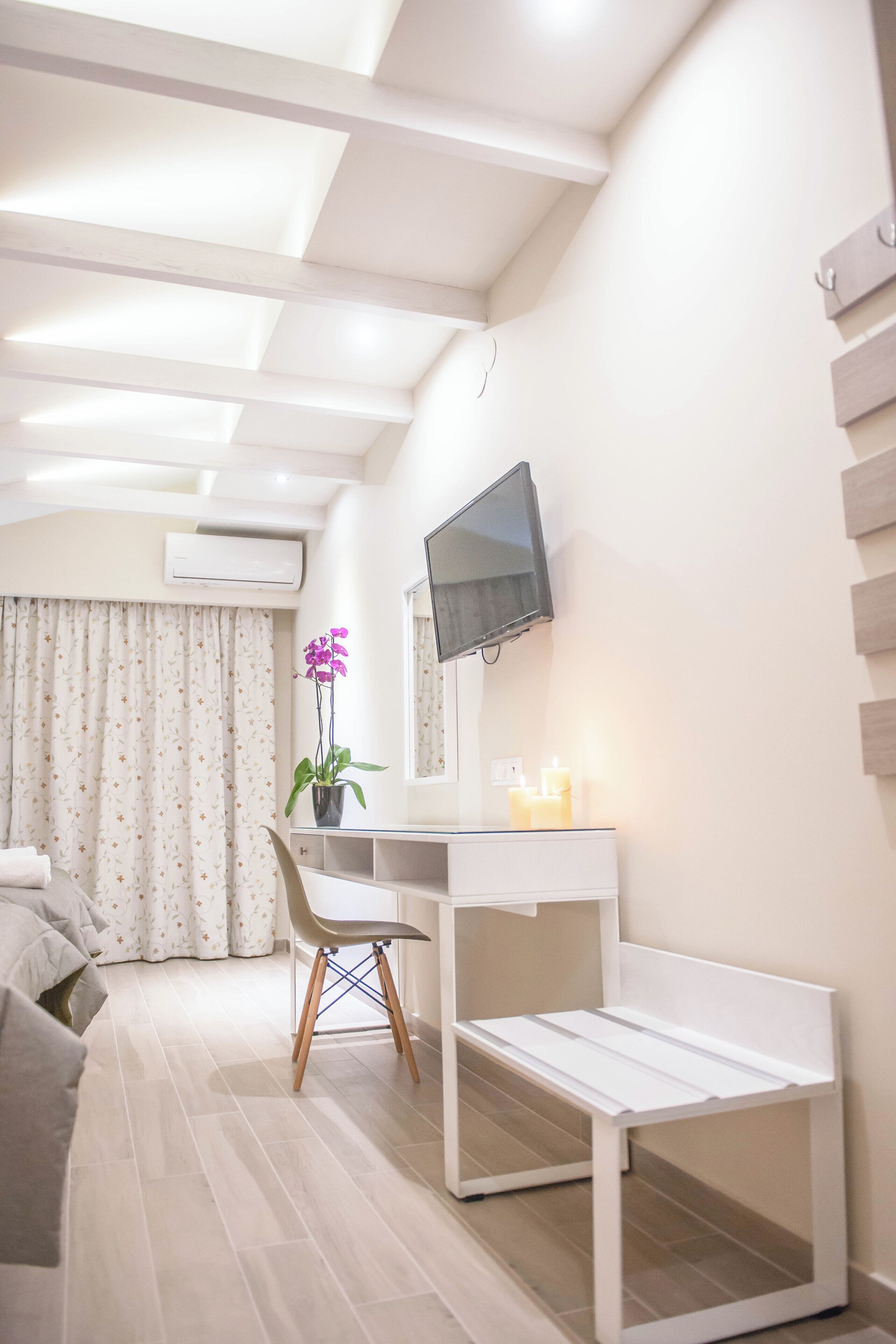 Une chambre moderne avec un bureau blanc, un téléviseur à écran plat, une orchidée en pot et un lit agrémenté d’un rideau à motifs.