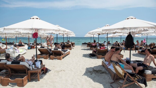 A beach with sun loungers and umbrellas, people relaxing, and the ocean in the background.