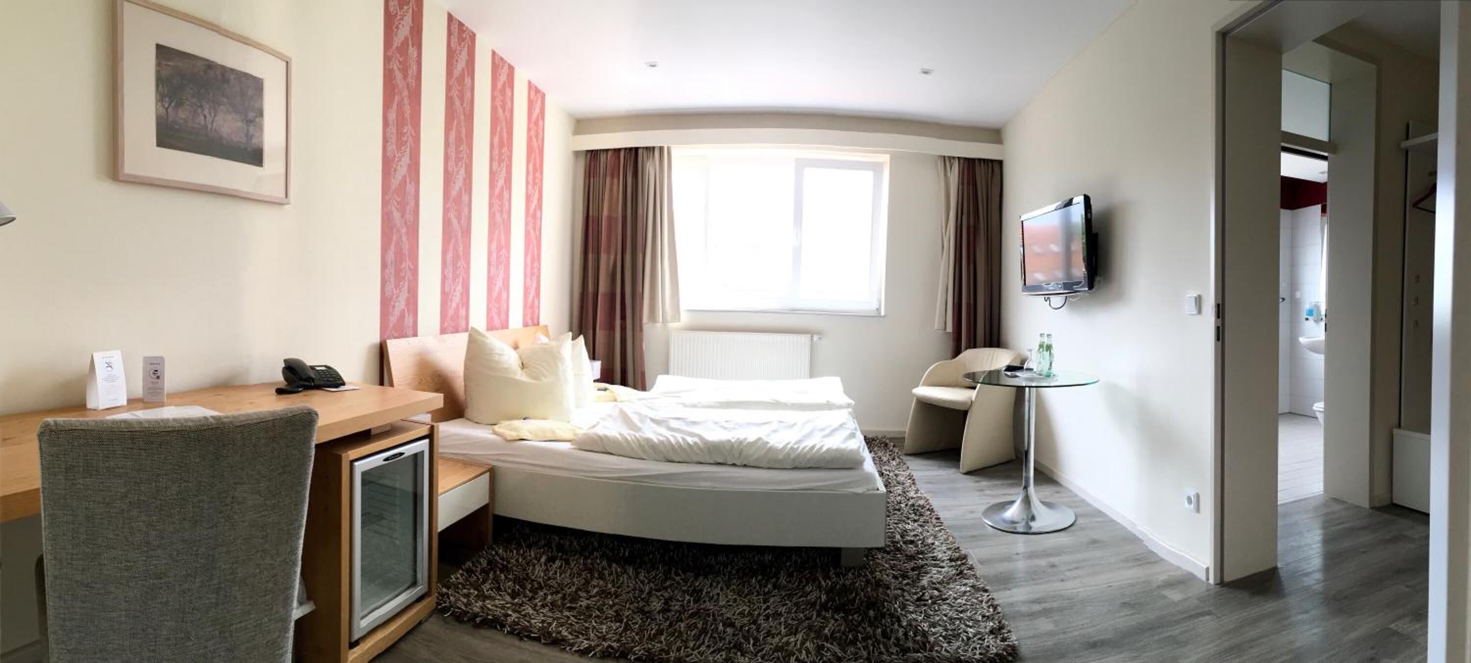 Ein Hotelzimmer mit Bett, Schreibtisch mit Stuhl, Fernseher und einem Fenster mit Vorhängen.