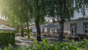 Un área de estar al aire libre con mesas y sillas bajo sombrillas, rodeada de árboles y un edificio de fachada de piedra.