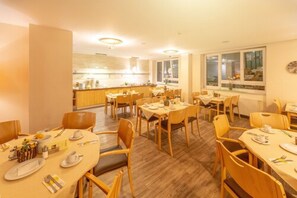 A dining area with multiple tables and chairs, a kitchen in the background, and large windows.