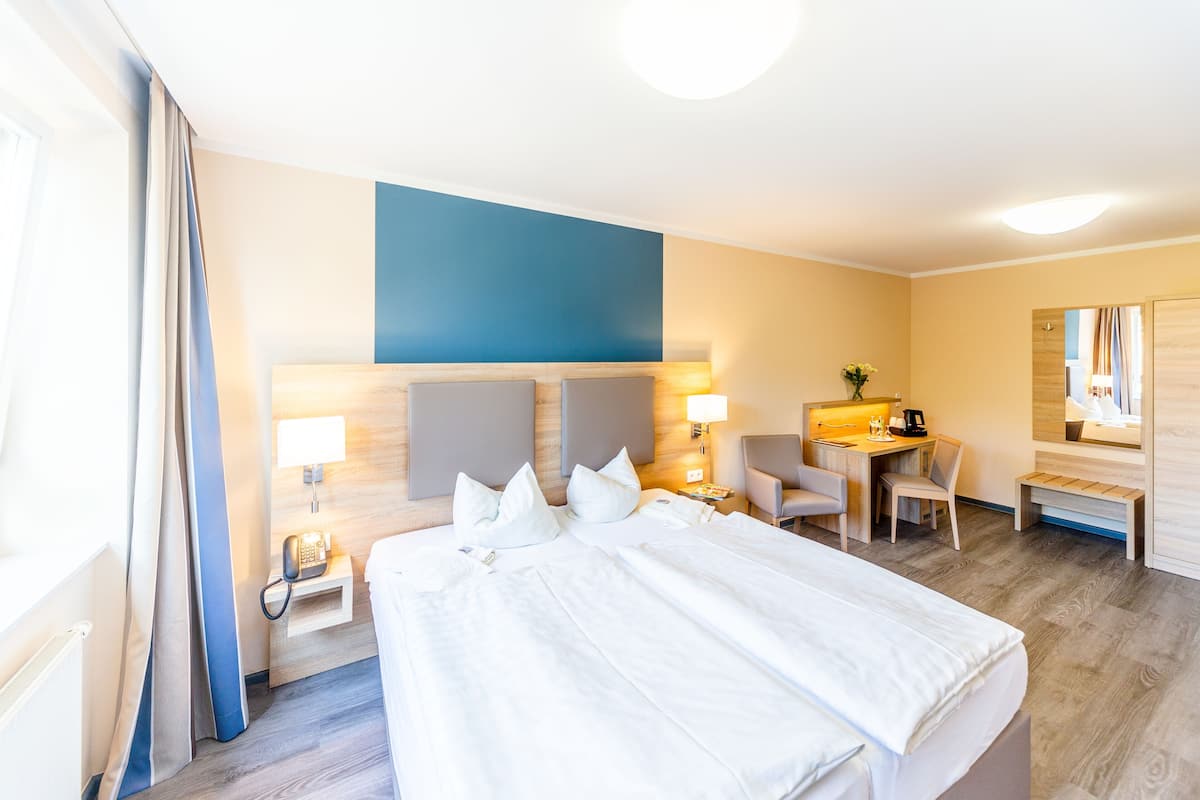 A hotel room with a large bed, bedside tables, a desk with a chair, a mirror, and a window with curtains.