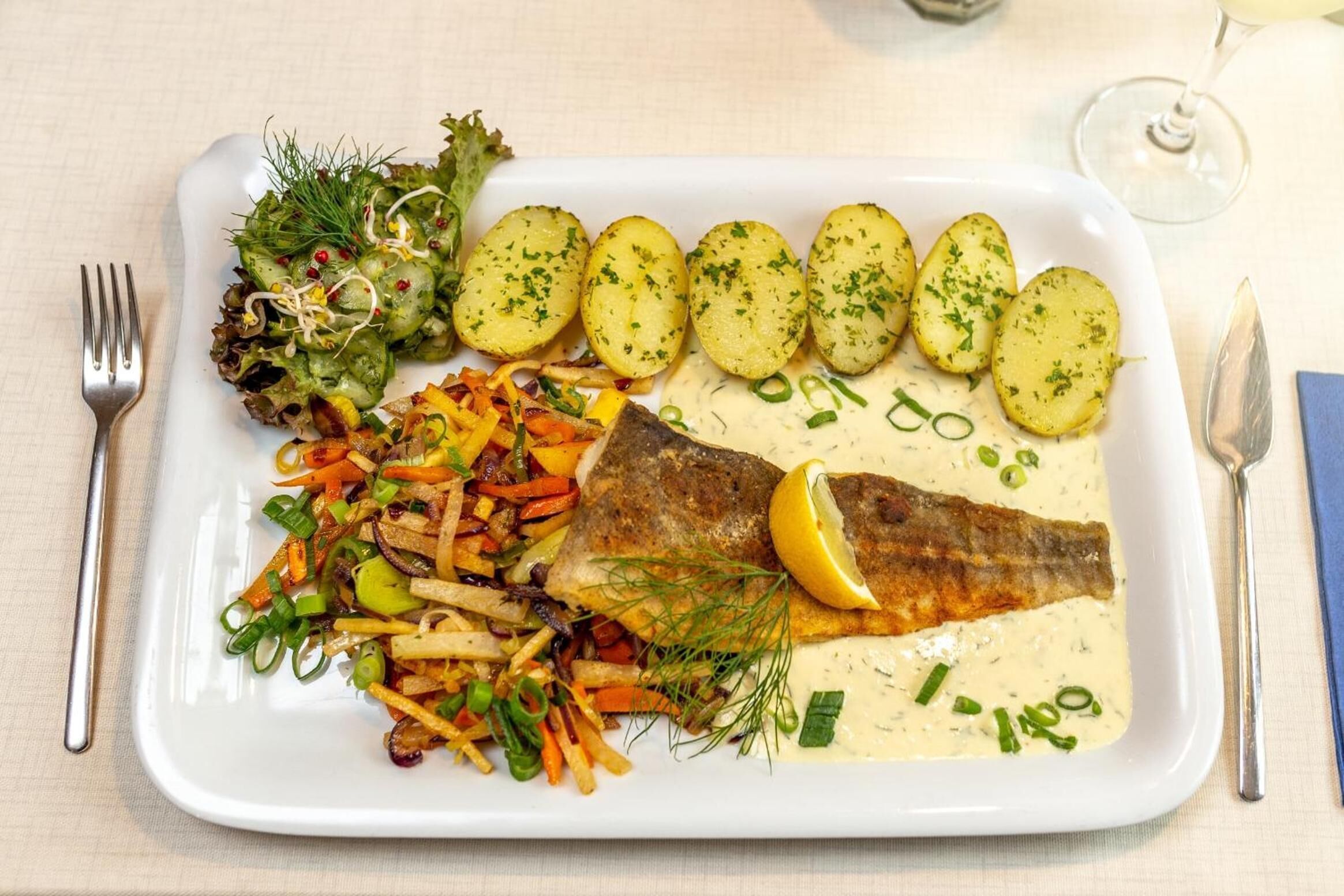 Un piatto bianco rettangolare con un filetto di pesce, patate bollite, un'insalata di verdure e patatine fritte.