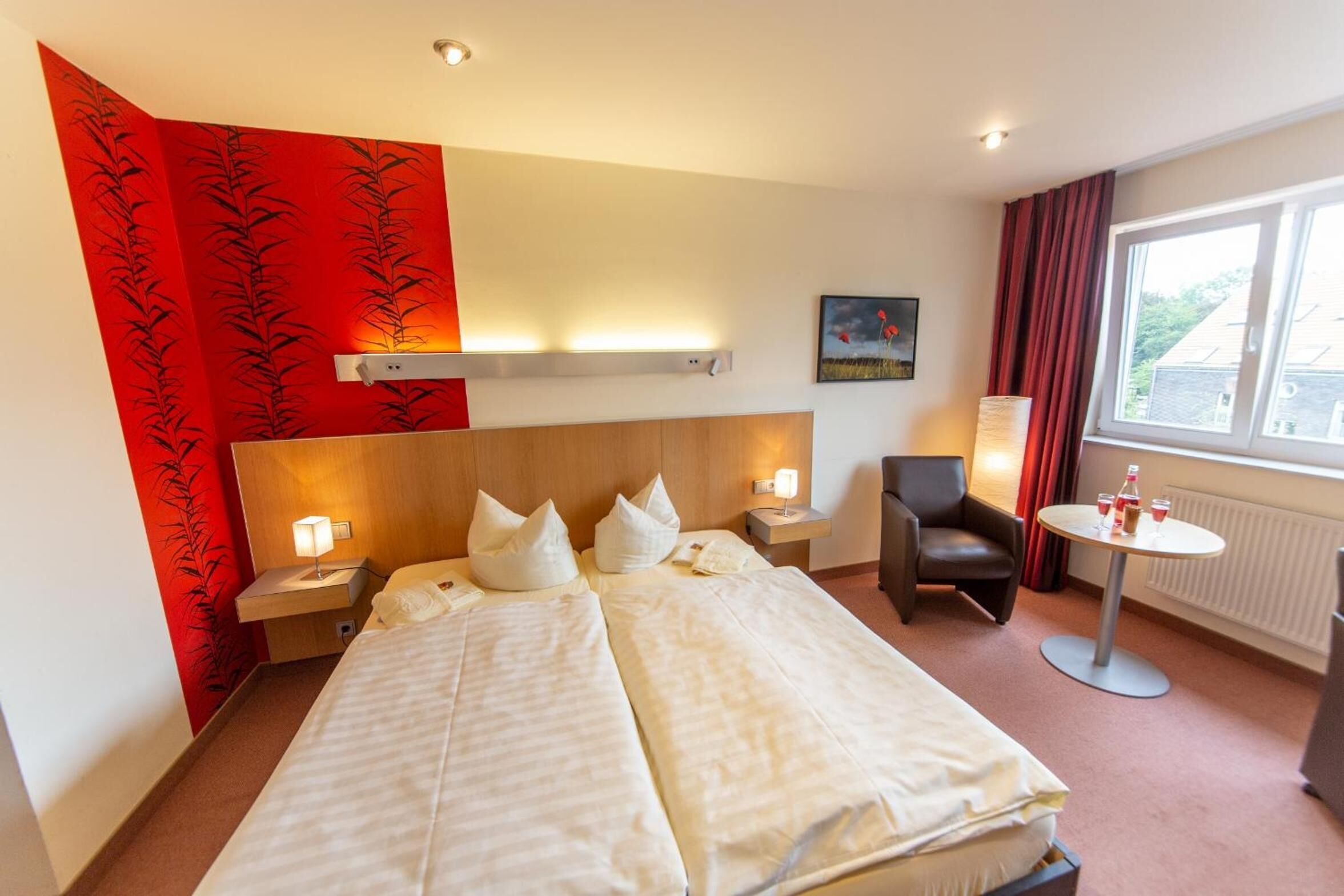 Une chambre d’hôtel avec un grand lit, une table de chevet avec une lampe, une chaise, une petite table avec des boissons et une fenêtre avec des rideaux.