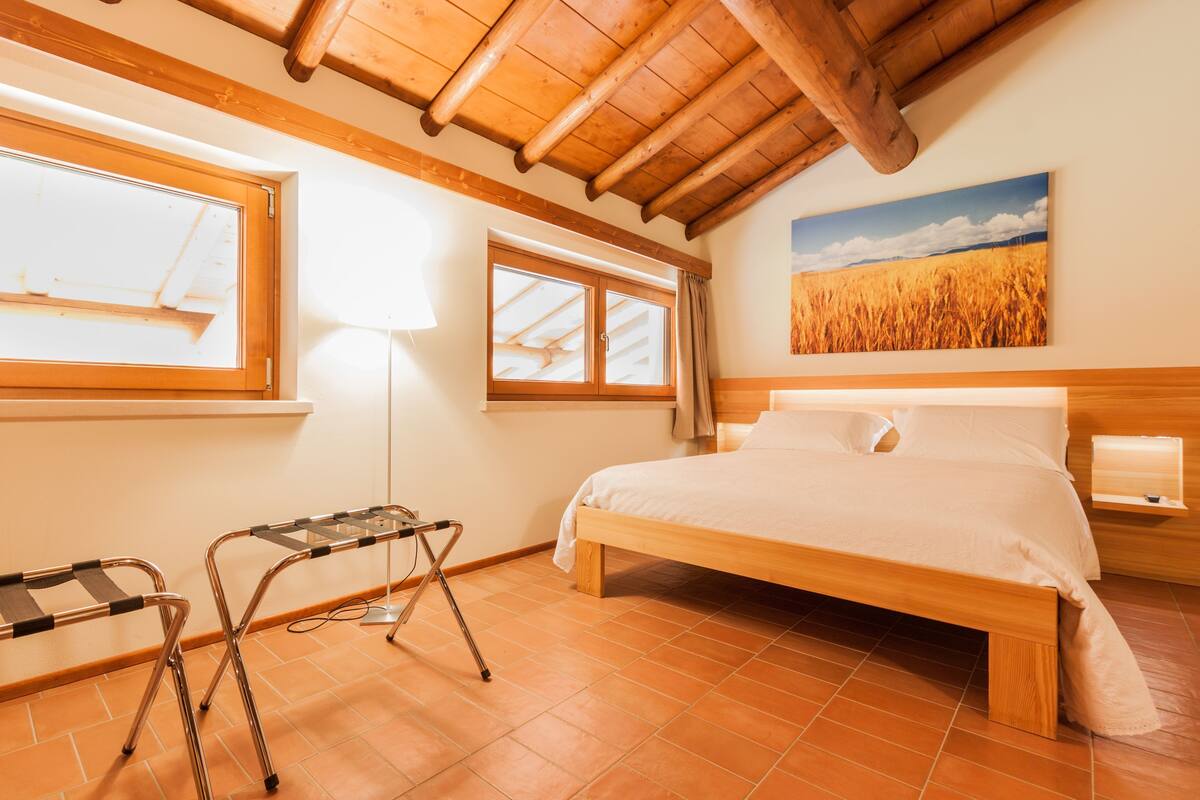 A bedroom with a wooden bed, two windows, a lamp, and a painting of a field.