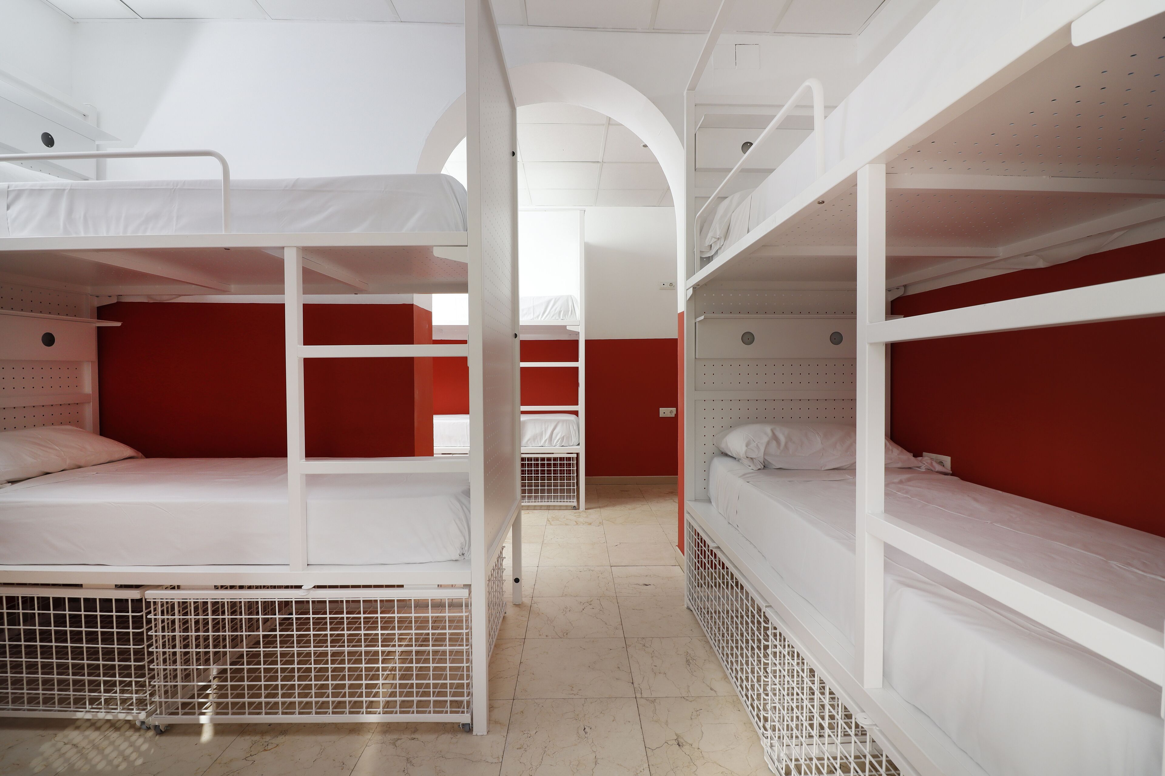 Un dormitorio con literas, camas blancas y paredes rojas, con suelo de baldosas y techo abovedado.
