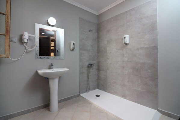 Un baño con lavamanos pedestal, espejo, secador de pelo portátil, cabezal de ducha manual y inodoro empotrado.