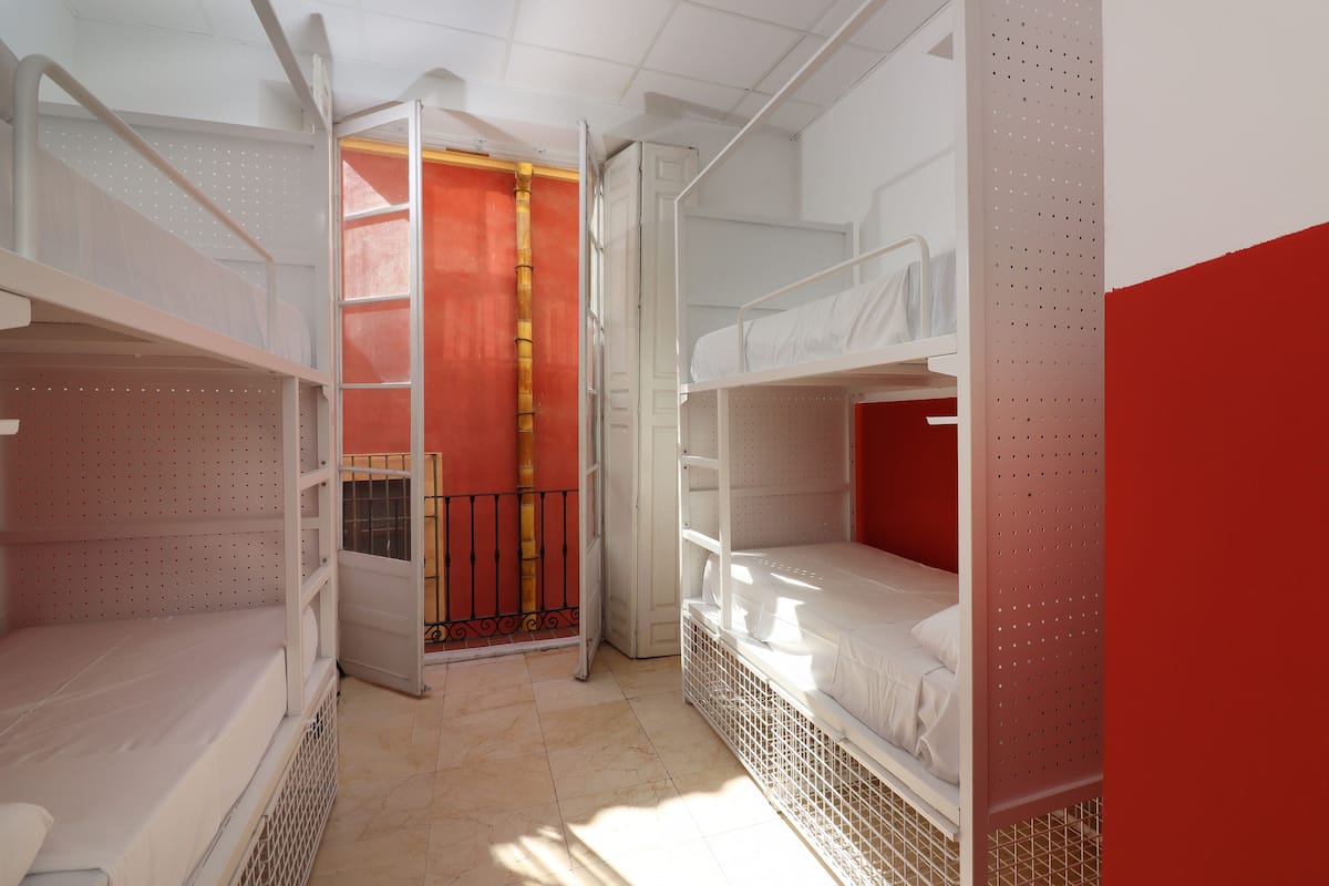 A dormitory room with bunk beds, a red door, and a window with a view.