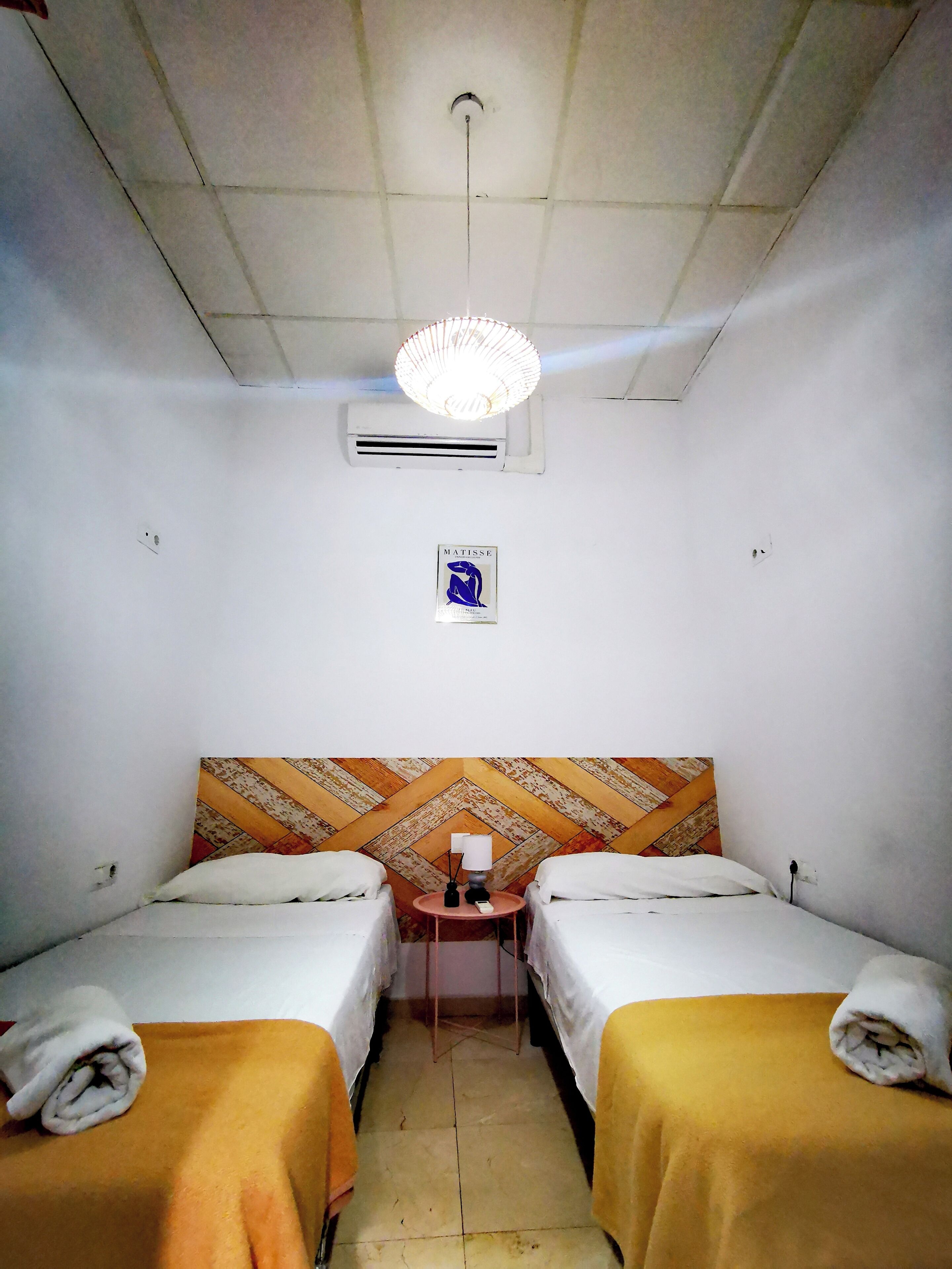 Een kleine hotelkamer met twee bedden, een bureau, een stoel, een bekeken hoofdbord, een airconditioning en een plafondlamp.