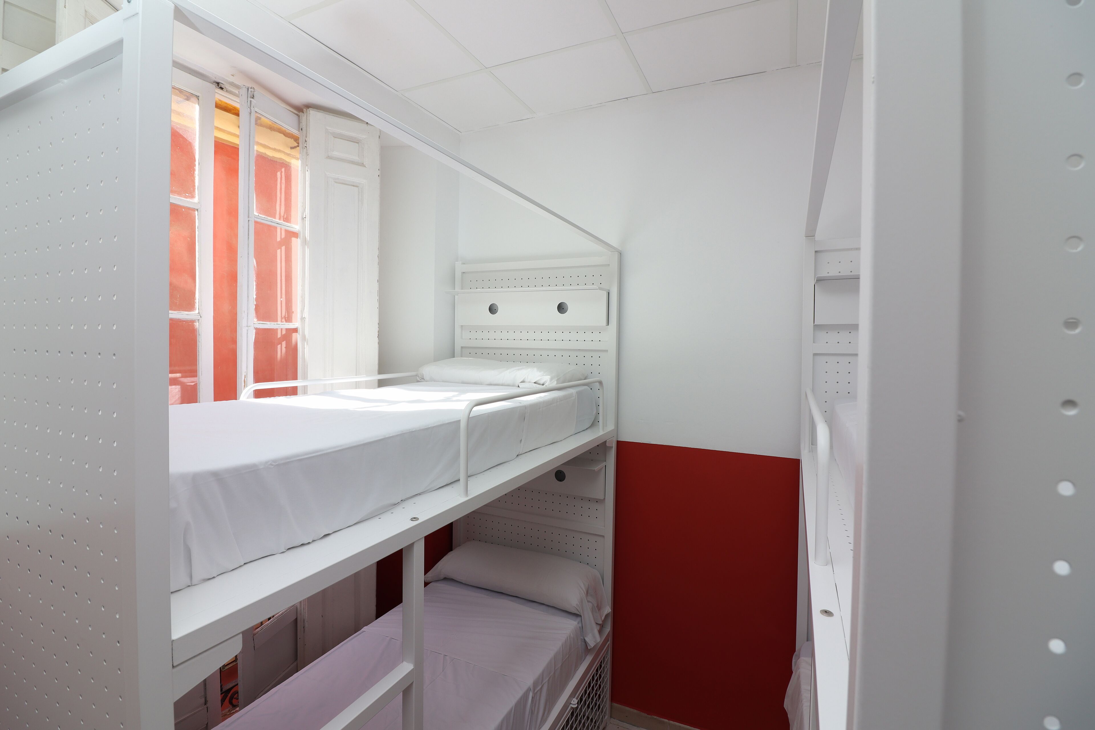 Een slaapkamer met stapelbed, raam, witte muren en een rode accentmuur.