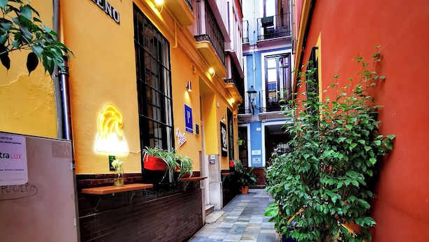 Un callejón estrecho con un edificio amarillo, un letrero de neón y plantas en macetas.