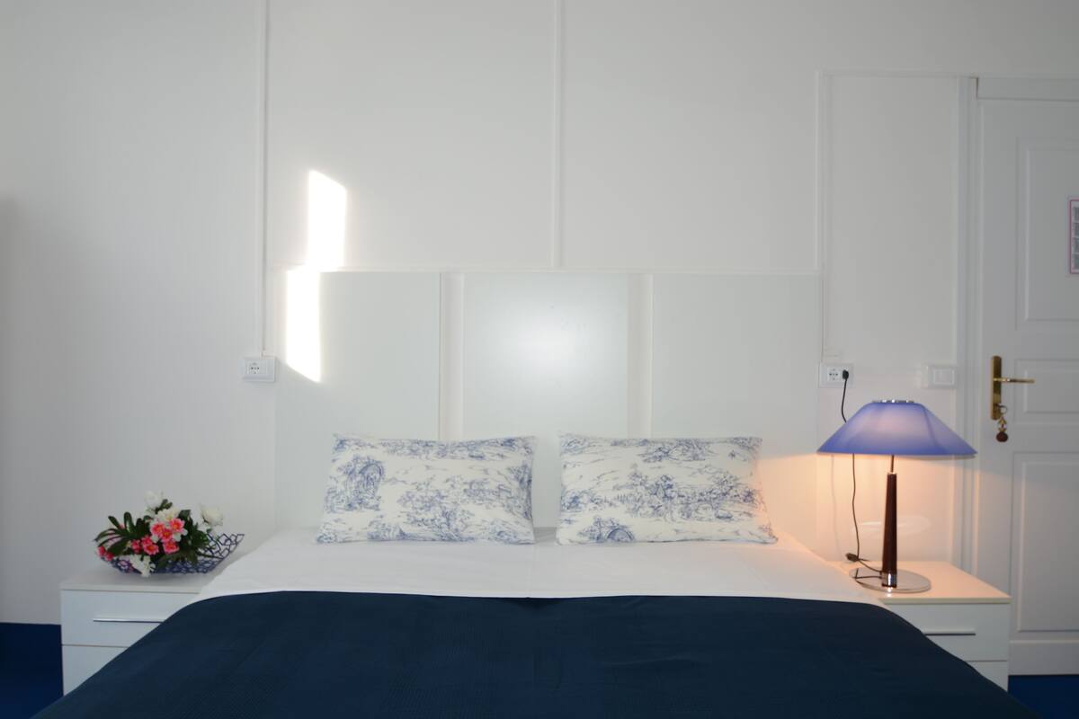 A bedroom with a bed, a nightstand with a lamp, and a wall-mounted shelf with a flower arrangement.