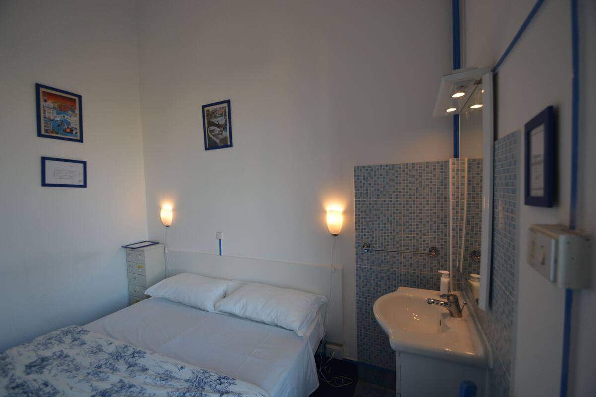 A bedroom with a bed, bedside lamps, a nightstand, and a bathroom with a sink and mirror.