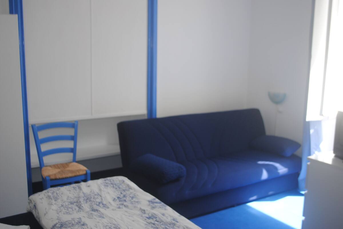 A room with a blue sofa, a small wooden chair, and a bed with a patterned bedspread.