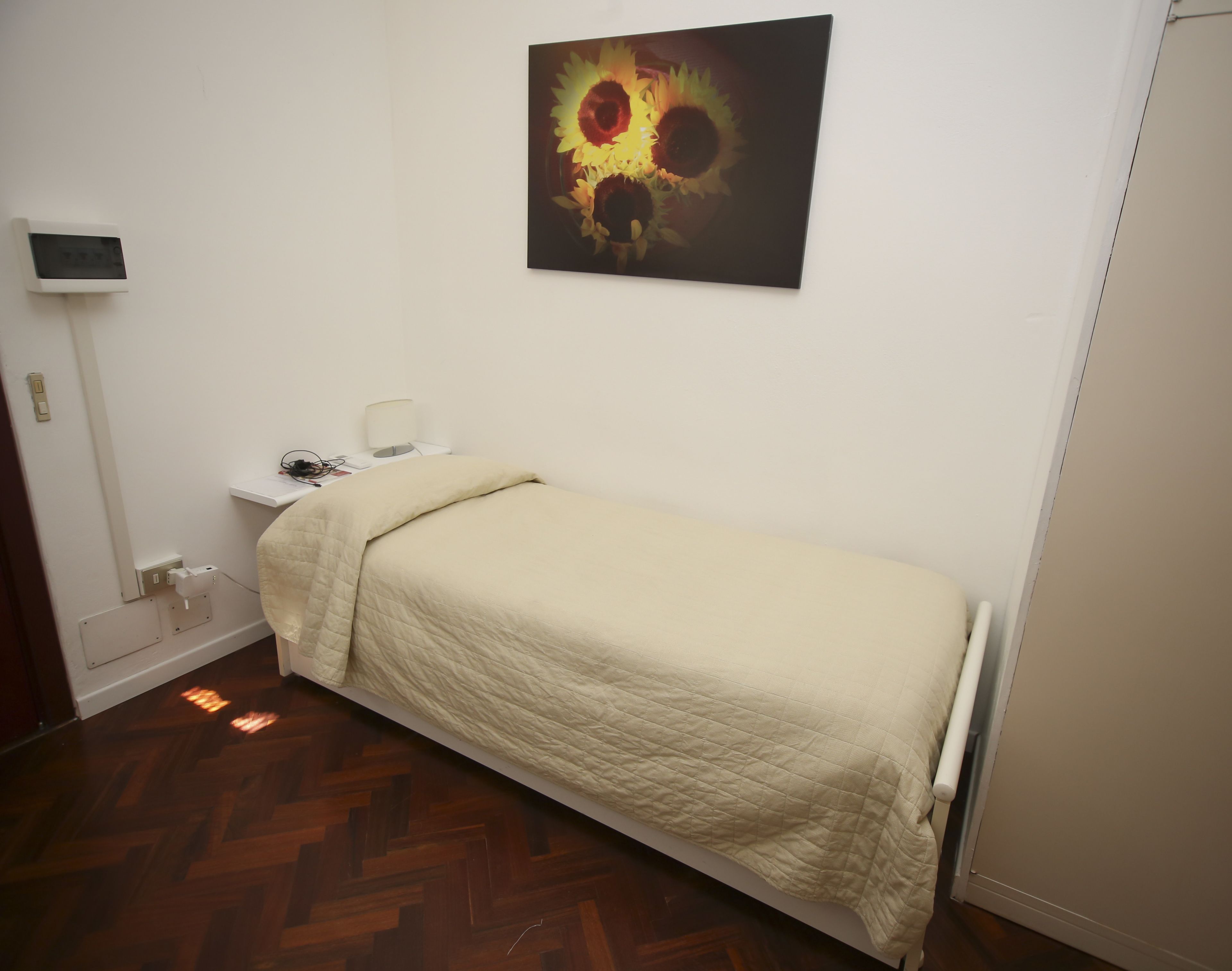 A small, neatly made room with a single bed, a bedside table with a lamp, and a wall-mounted thermostat.