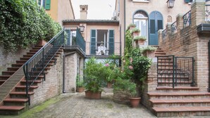 Un patio estrecho con una escalera de piedra que lleva a un balcón en el segundo piso, rodeado de macetas con plantas y un muro de ladrillo.